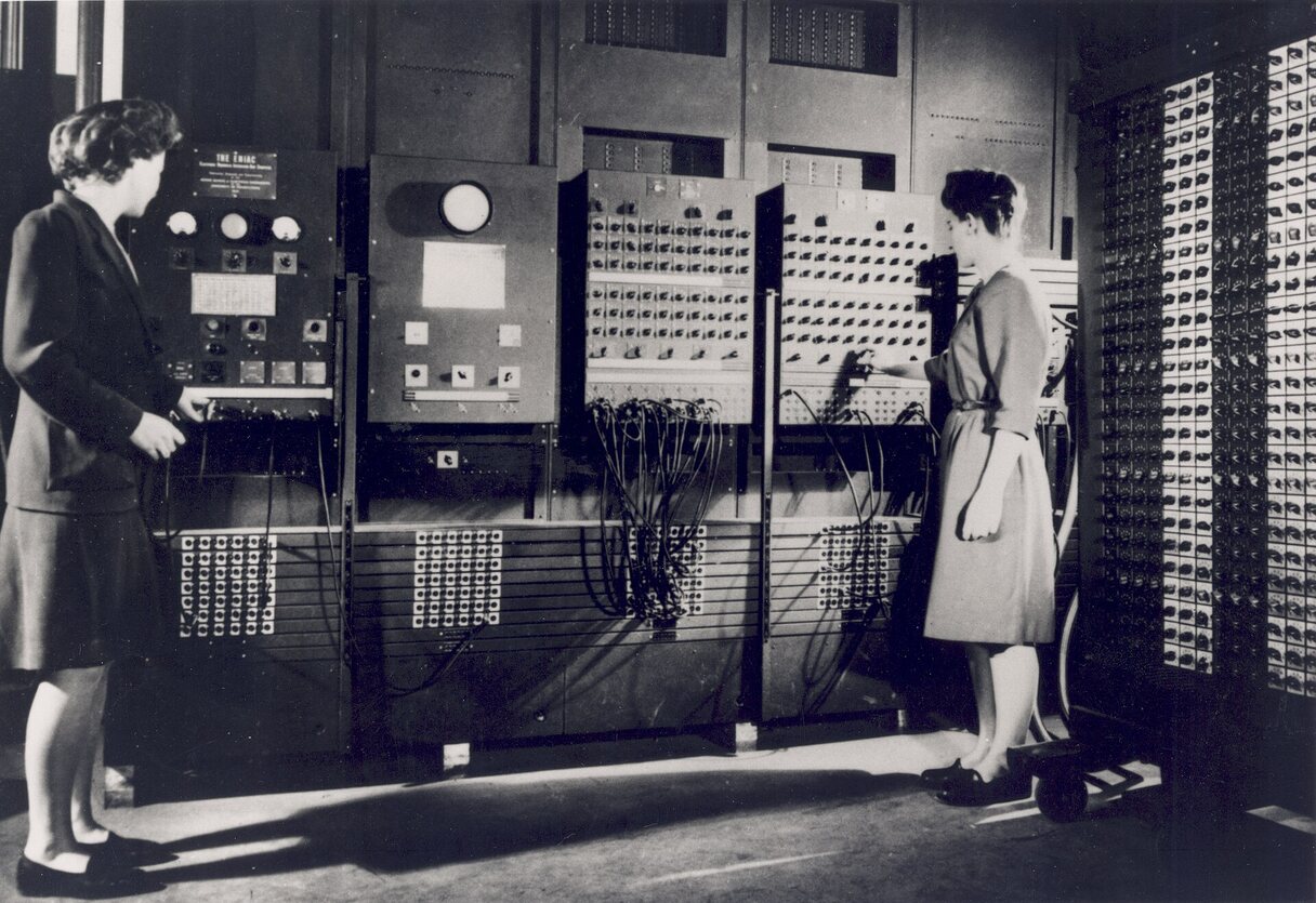 Two women standing at ENIAC’s panels of switches and cables, operating the machine.