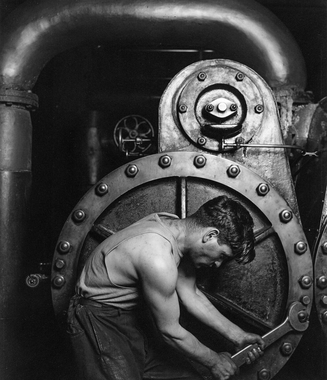 A power house mechanic bent over a steam pump, wrench in both hands. Photograph by Lewis Hine, 1920.