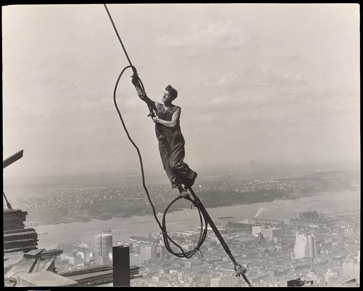 An ironworker balancing on a cable high above the city, slinging a hoisting line.