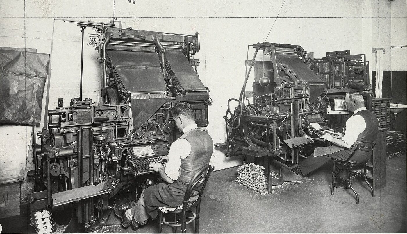 Operators seated at rows of linotype machines, setting perfect type.
