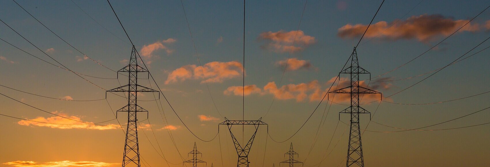 Rows of high-voltage transmission towers carrying power lines toward the horizon at sunset. Photograph by Matthew Henry.
